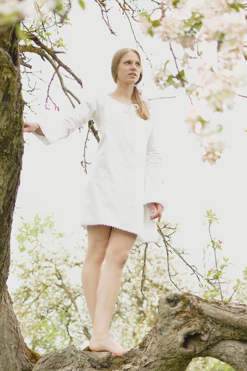 Pode incluir: Uma jovem com um vestido branco est&aacute; em um galho de &aacute;rvore em um pomar florido. Os galhos est&atilde;o cobertos de flores brancas.