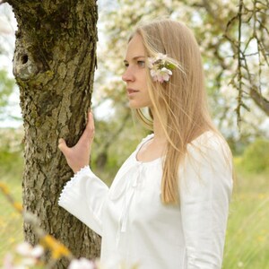 Pode incluir: Uma jovem com cabelo comprido e loiro est&aacute; em um campo de &aacute;rvores floridas. Ela est&aacute; vestindo uma blusa branca e tem uma flor atr&aacute;s da orelha.