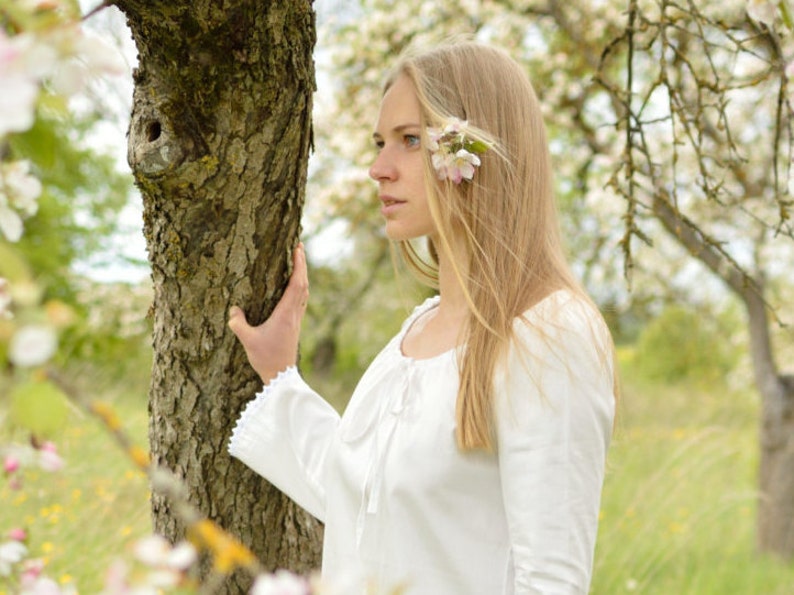 Pode incluir: Uma jovem com cabelo comprido e loiro est&aacute; em um campo de &aacute;rvores floridas. Ela est&aacute; vestindo uma blusa branca e tem uma flor atr&aacute;s da orelha.