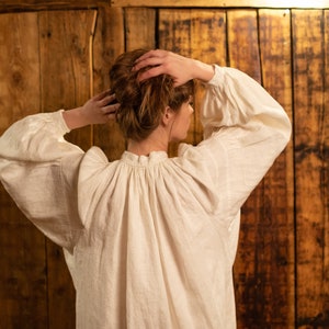 May include: A white linen blouse with a gathered neckline and long sleeves. The blouse is worn by a person with brown hair, standing in front of a wooden wall.