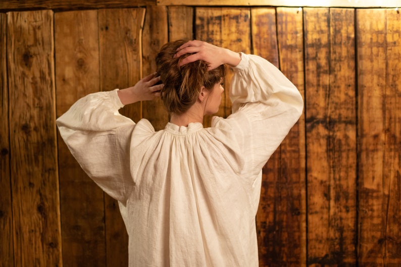 May include: A white linen blouse with a gathered neckline and long sleeves. The blouse is worn by a person with brown hair, standing in front of a wooden wall.