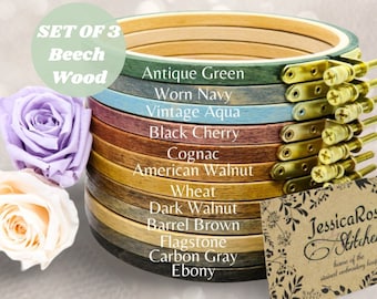 May include: Set of 3 beech wood embroidery hoops in various stained wood finishes, including antique green, worn navy, vintage aqua, black cherry, cognac, American walnut, wheat, dark walnut, barrel brown, flagstone, carbon gray, and ebony. The hoops are stacked on top of each other and are labeled with their respective finishes. The image also includes a small sign that reads "Jessica Rose Stitchery, home of the stained embroidery hoops."
