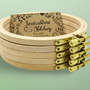 May include: A set of six wooden embroidery hoops with brass screw-on clamps. The hoops are stacked on top of each other and a business card with the text "JessicaRose Stitchery home of the hand embroidery hoop" is visible in the background.