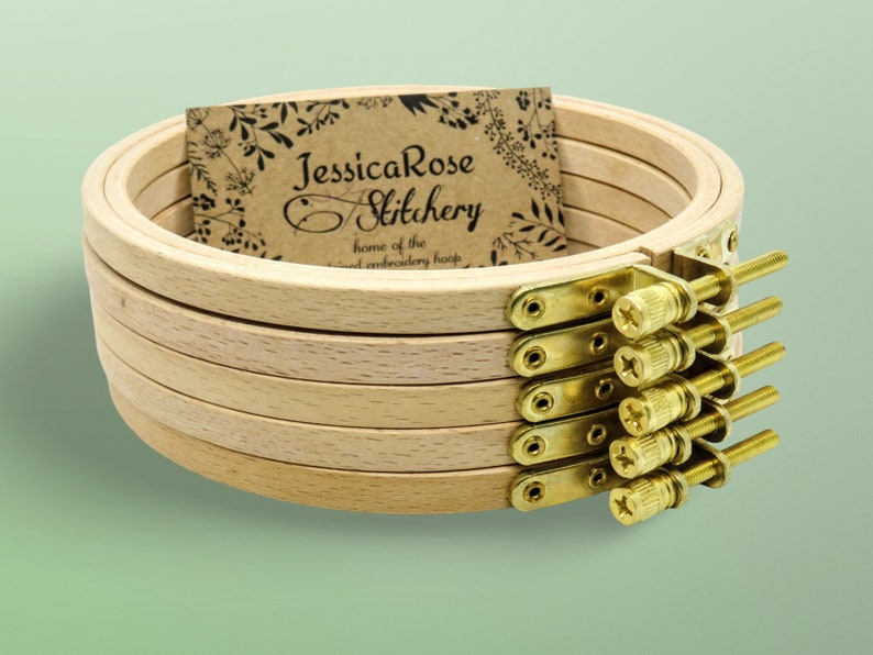 May include: A set of six wooden embroidery hoops with brass screw-on clamps. The hoops are stacked on top of each other and a business card with the text "JessicaRose Stitchery home of the hand embroidery hoop" is visible in the background.