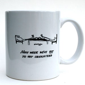 Puede incluir: Taza de café de cerámica blanca con una ilustración de dibujos animados en blanco y negro de dos personas sentadas en una mesa. El texto "Next week we've got to get organized" está impreso debajo de la ilustración.