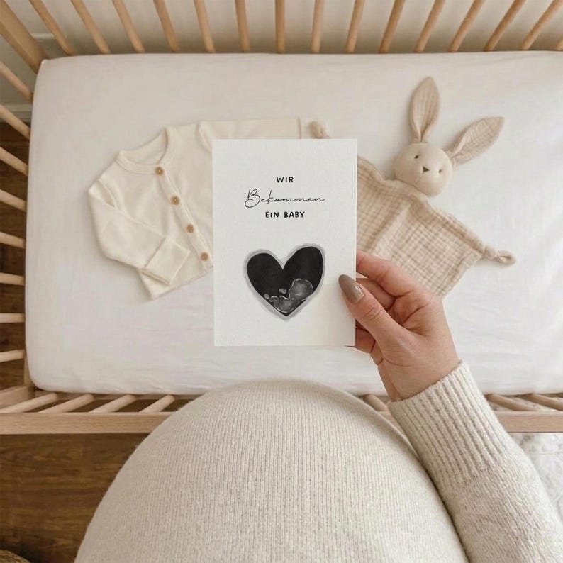 K&ouml;nnte beinhalten: Eine Schwangerschaftsank&uuml;ndigungskarte mit dem Text "WIR Bekommen EIN BABY" und einem herzf&ouml;rmigen Ultraschallbild. Die Karte wird vor dem Bauch einer schwangeren Person gehalten. Ein cremefarbener Baby-Cardigan und ein Stoffhase befinden sich im Hintergrund.