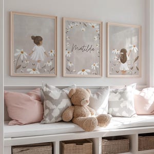 May include: Three framed art prints featuring illustrations of a girl in a white dress among daisies. The middle print has the name "Matilda" in script. The prints are displayed above a window seat with pillows and a teddy bear.