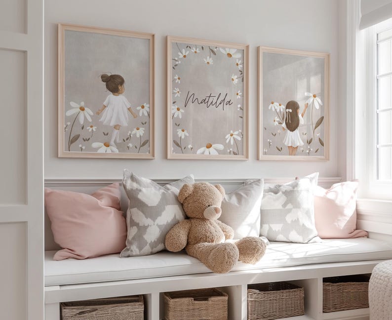 May include: Three framed art prints featuring illustrations of a girl in a white dress among daisies. The middle print has the name "Matilda" in script. The prints are displayed above a window seat with pillows and a teddy bear.