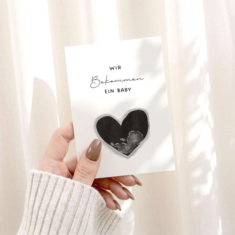 K&ouml;nnte beinhalten: Eine wei&szlig;e Karte mit dem Text "WIR Bekommen EIN BABY" und einem schwarzen Herz mit einem Aquarellbild eines F&ouml;tus. Die Karte wird vor einem weichen, neutralen Hintergrund gehalten.