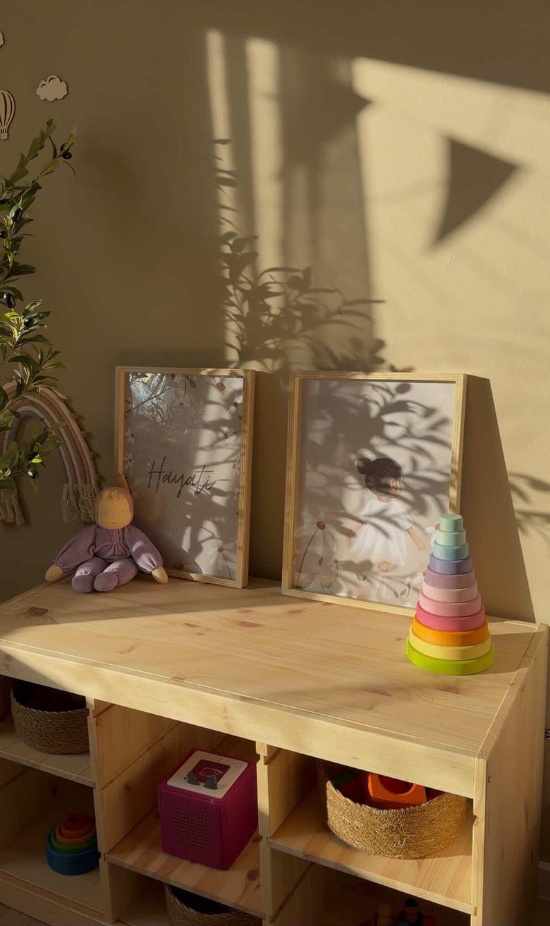 May include: A wooden framed print with the word "Hayat" in cursive script, a daisy print, and a colorful wooden stacking toy sit on a wooden table. A purple doll and a rainbow decoration are also on the table.
