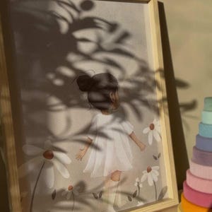 May include: A framed print featuring a young girl in a white dress amidst daisies. The artwork has a soft, pastel color palette and is set in a light wooden frame. A colorful wooden stacking toy is visible on the right side of the frame.