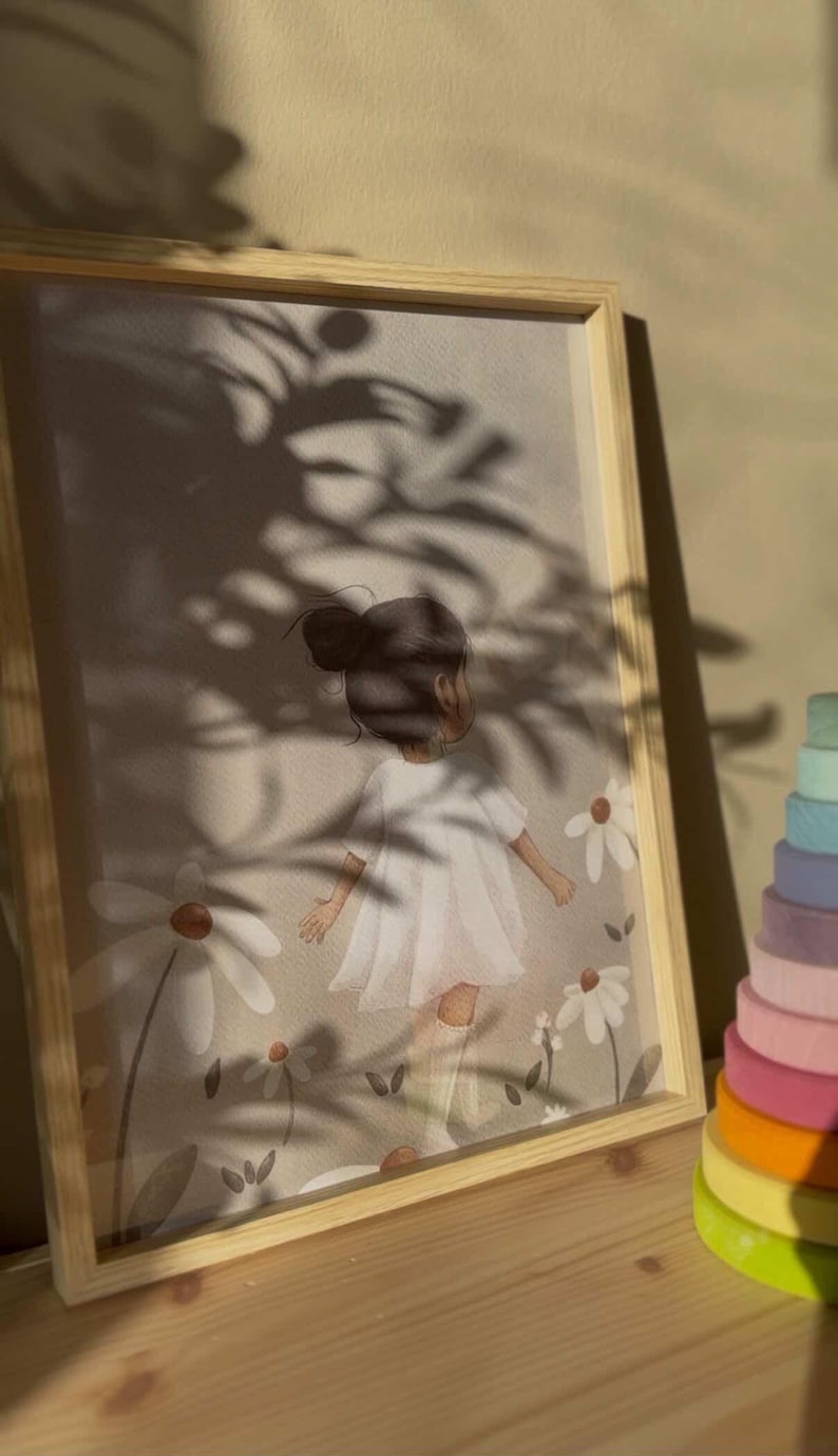 May include: A framed print featuring a young girl in a white dress amidst daisies. The artwork has a soft, pastel color palette and is set in a light wooden frame. A colorful wooden stacking toy is visible on the right side of the frame.