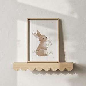 May include: A framed watercolor print featuring a light brown bunny holding daisies. The artwork has a soft, pastel color palette with a dotted background. The frame is light wood, and the print is displayed on a scalloped wooden shelf.