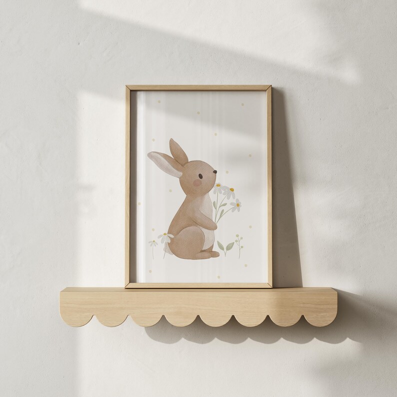 May include: A framed watercolor print featuring a light brown bunny holding daisies. The artwork has a soft, pastel color palette with a dotted background. The frame is light wood, and the print is displayed on a scalloped wooden shelf.