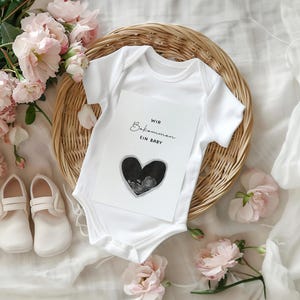 K&ouml;nnte beinhalten: Ein wei&szlig;er Baby-Body mit einer Karte mit der Aufschrift "Wir Bekommen Ein Baby" und einem herzf&ouml;rmigen Ultraschallbild. Der Body liegt auf einem Weidenkorb, umgeben von rosa Blumen und einem Paar wei&szlig;er Babyschuhe.