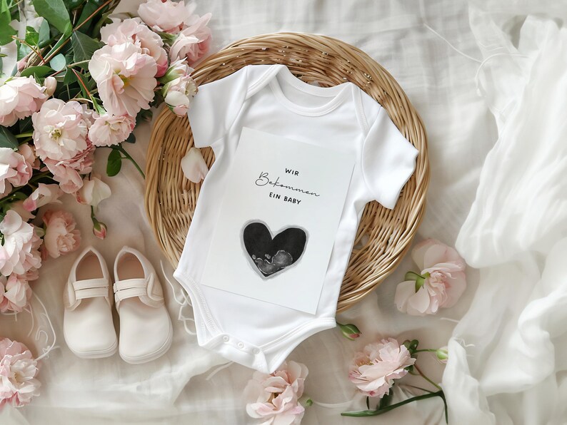 K&ouml;nnte beinhalten: Ein wei&szlig;er Baby-Body mit einer Karte mit der Aufschrift "Wir Bekommen Ein Baby" und einem herzf&ouml;rmigen Ultraschallbild. Der Body liegt auf einem Weidenkorb, umgeben von rosa Blumen und einem Paar wei&szlig;er Babyschuhe.