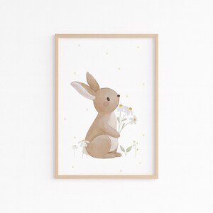 May include: Watercolor illustration of a tan bunny holding white daisies, set against a white background with yellow polka dots. The artwork is framed in a light brown wooden frame, perfect for nursery decor.