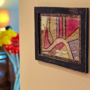 May include: A framed vintage map of New Orleans, printed in shades of pink, yellow, and blue, with a brown river winding through the city. The frame is black with a distressed finish.