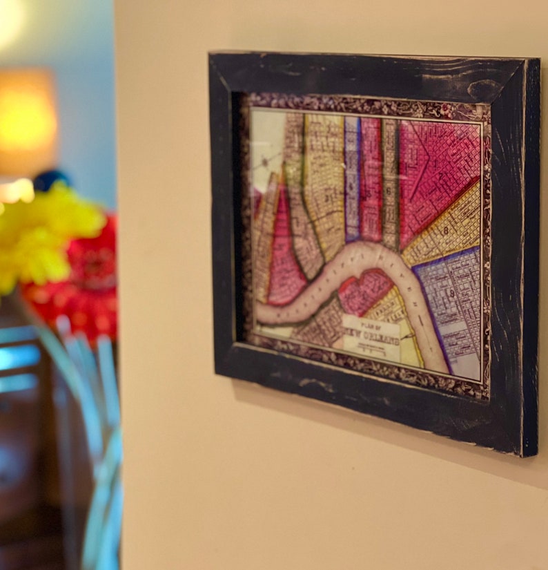 May include: A framed vintage map of New Orleans, printed in shades of pink, yellow, and blue, with a brown river winding through the city. The frame is black with a distressed finish.