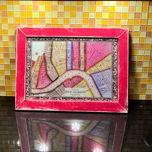 May include: A vintage map of New Orleans in a pink wood frame. The map is printed on paper and shows the streets and blocks of the city. The map is titled "Plan of New Orleans".