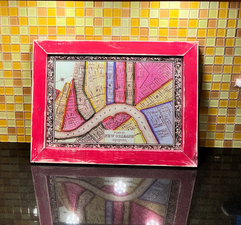 May include: A vintage map of New Orleans in a pink wood frame. The map is printed on paper and shows the streets and blocks of the city. The map is titled "Plan of New Orleans".