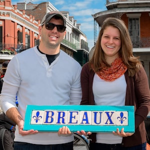 May include: A couple poses in front of a building with a sign that reads "BREAUX" in blue letters on a turquoise background. The sign has fleur-de-lis designs on either end.