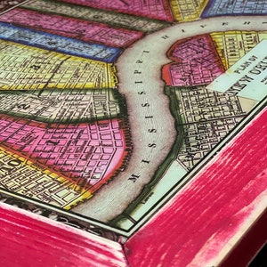 May include: A framed vintage map of New Orleans, Louisiana, with a pink wood frame. The map shows the city's streets and the Mississippi River. The title of the map is "Plan of New Orleans".
