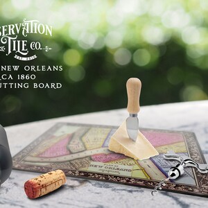 May include: A glass cutting board featuring a vintage map of New Orleans, circa 1860. The board is surrounded by a decorative border and includes the text "Preservation Tile Co.". The image also shows a cheese knife, a corkscrew, and a wine bottle.