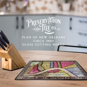 May include: A glass cutting board featuring a vintage map of New Orleans circa 1860. The board is surrounded by a black border and has a decorative design. The text "Preservation Tile Co." is printed in white on the board. The board is sitting on a wooden table with a set of knives in a wooden block.