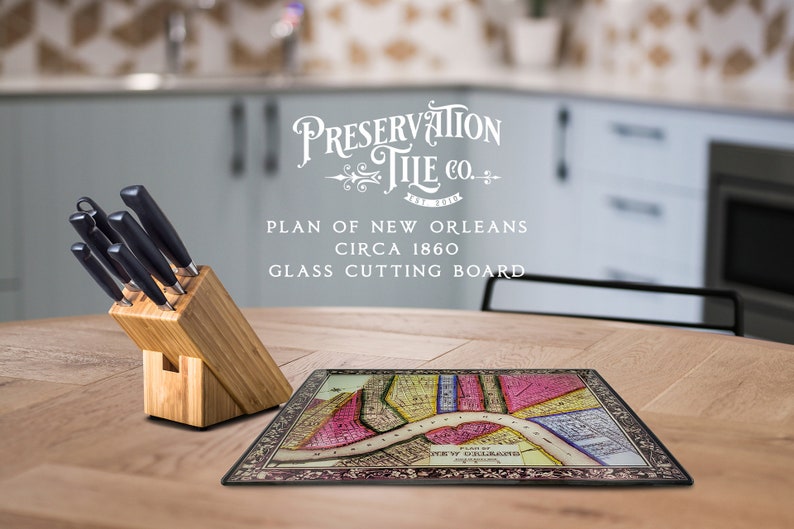 May include: A glass cutting board featuring a vintage map of New Orleans circa 1860. The board is surrounded by a black border and has a decorative design. The text "Preservation Tile Co." is printed in white on the board. The board is sitting on a wooden table with a set of knives in a wooden block.