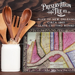 May include: A glass cutting board featuring a vintage map of New Orleans, circa 1860. The board has a dark brown frame with floral details and the text "Preservation Tile Co. Est 2010" and "Plan of New Orleans Circa 1860 Glass Cutting Board".