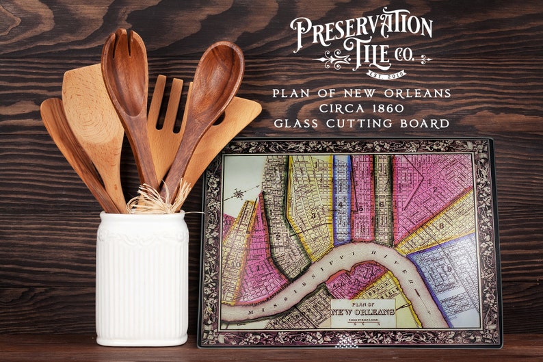 May include: A glass cutting board featuring a vintage map of New Orleans, circa 1860. The board has a dark brown frame with floral details and the text "Preservation Tile Co. Est 2010" and "Plan of New Orleans Circa 1860 Glass Cutting Board".