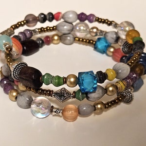 May include: A multi-strand wrap bracelet with a variety of beads in shades of gray, brown, blue, and gold. The beads include faceted cubes, round pearls, and wooden elements. The bracelet is approximately 7 inches in circumference.