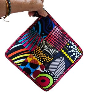 May include: A colorful, square quilted potholder with a vibrant patchwork design. The potholder features a variety of patterns and colors, including red, blue, yellow, and black. It has a red border and a small loop for hanging.