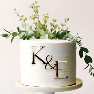 Könnte beinhalten: Weiße Hochzeitstorte mit goldenen Buchstaben, die "K&E" auf der Oberseite buchstabieren, verziert mit grünen und weißen Blumen.