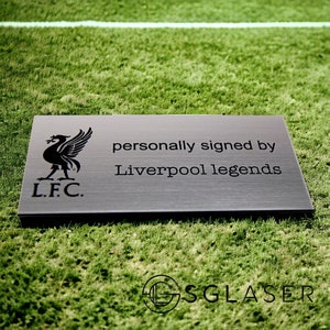 Könnte beinhalten: Eine schwarze Metallplakette mit dem Wappen des Liverpool Football Club und dem eingravierten Text "persönlich signiert von Liverpool-Legenden".