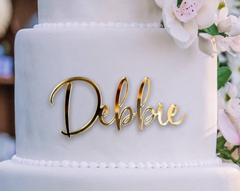 Può includere: Una torta nuziale bianca con un topper in acrilico a specchio dorato che dice "Debbie".