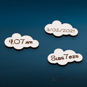 Puede incluir: Tres adornos de madera blanca en forma de nube con el texto "3/02/2021", "4:07 AM" y "8 lbs 7 ozs" escrito en tinta marrón.