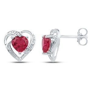 May include: A pair of silver heart-shaped earrings with a red heart-shaped gemstone in the center of each earring. The heart is surrounded by a silver halo with smaller clear gemstones.