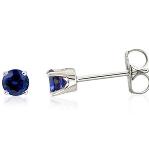 May include: A pair of stud earrings featuring round, deep blue gemstones set in a silver metal. One earring is shown separately, and the other is shown with the post and backing. The earrings are displayed against a white background.