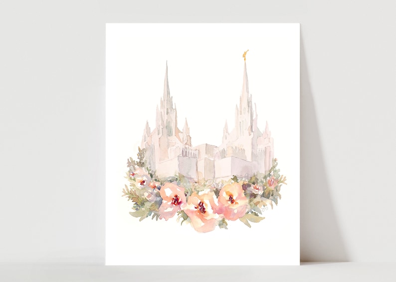 May include: Watercolor print of a white building with two spires, topped with a gold accent. The building is surrounded by a wreath of pink and peach flowers and green foliage, set against a white background.