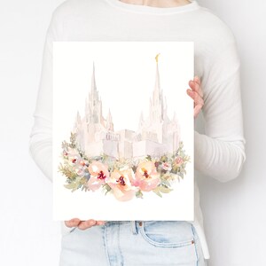 May include: Watercolor print of a white building with tall spires, possibly a temple, adorned with a floral wreath of peach and pink flowers. The artwork is held by a person.