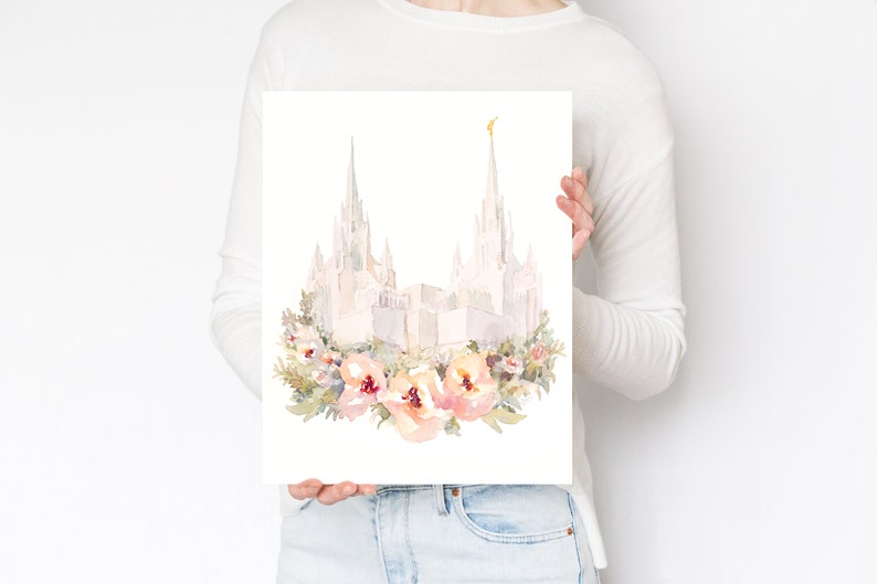 May include: Watercolor print of a white building with tall spires, possibly a temple, adorned with a floral wreath of peach and pink flowers. The artwork is held by a person.