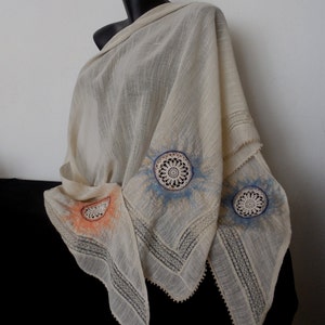 Può includere: Sciarpa color crema con bordi in pizzo intricati e disegni circolari colorati. La sciarpa è drappeggiata su un manichino nero, mettendo in risalto la sua delicata consistenza e gli abbellimenti artistici. I disegni includono elementi arancioni, blu e bianchi.
