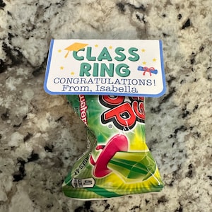 May include: A blue and white card with the text "CLASS RING CONGRATULATIONS! From, Isabella" attached to a green and yellow Pop Rocks candy package.
