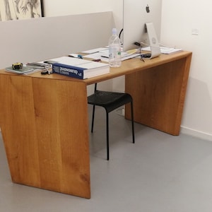 May include: A light wood desk with a slightly angled edge. The desk has a book, a water bottle, and a computer monitor on top. A black chair sits in front of the desk.