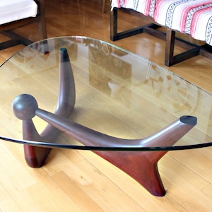 Könnte beinhalten: Ein moderner Glas-Couchtisch aus der Mitte des Jahrhunderts mit einem einzigartigen, skulpturalen Holzsockel. Der Sockel besteht aus drei geschwungenen Beinen, die sich in der Mitte treffen und eine unverwechselbare Form bilden. Die Glasplatte ist klar und oval.