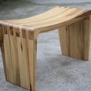 Puede incluir: Un taburete de madera con un asiento curvo y un diseño único. El taburete está hecho de madera clara con un acabado natural.