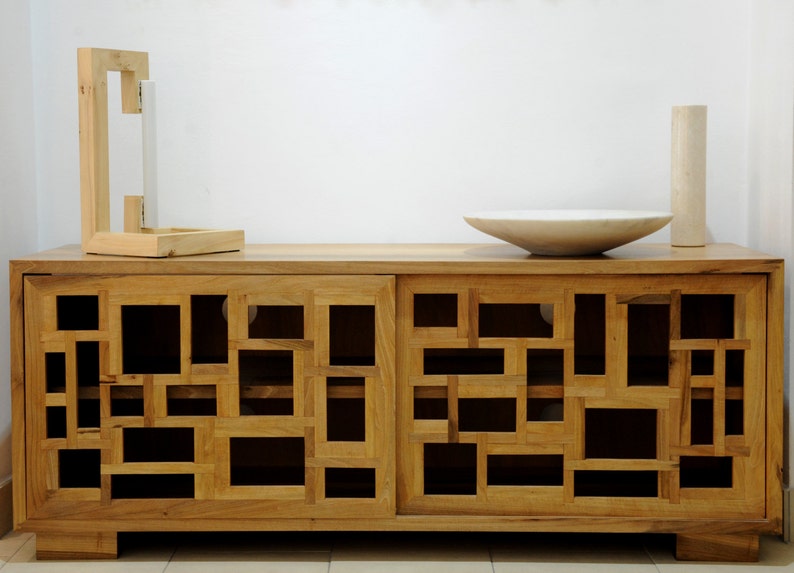 May include: A wooden cabinet with two doors featuring a geometric pattern of square cutouts. The cabinet has a light brown finish and sits on a white surface.
