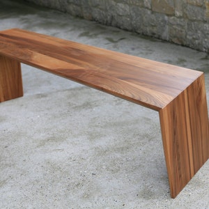 Puede incluir: Banco de madera moderno con acabado natural. El banco tiene un asiento rectangular y patas anguladas, creando un diseño elegante. La veta de la madera es visible, mostrando la belleza natural. El banco mide aproximadamente 152 cm de largo.
